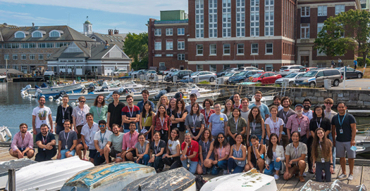 group photo of BMM summer school participants standing on a dock, behind 4 upturned boats